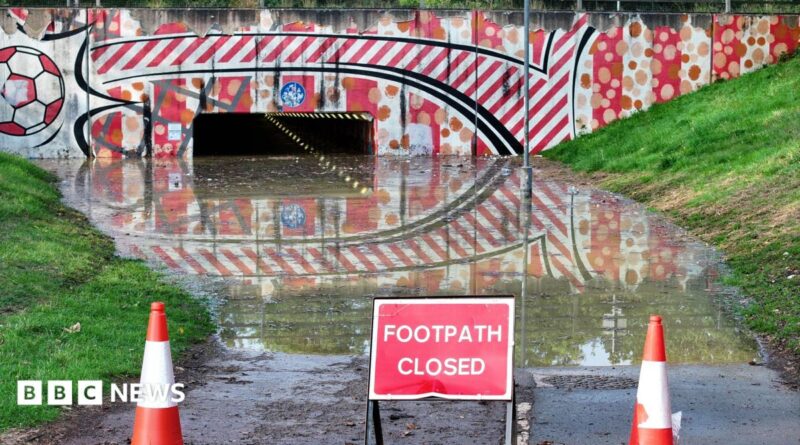 A flooded footpath leading to an underpath with warning cones and a "footpath closed" sign