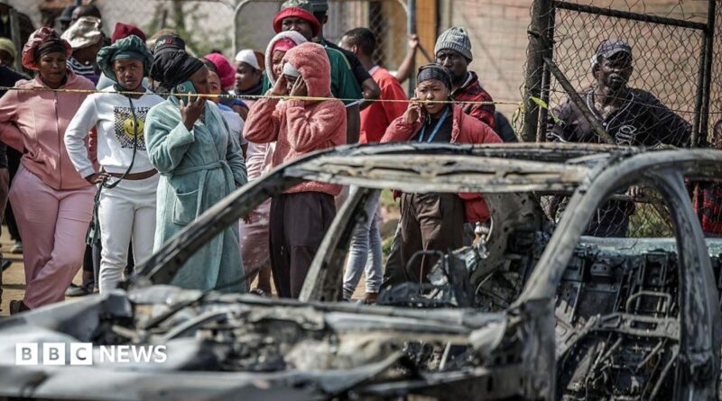 A crowd of people look on close to a burned out car