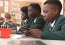 Students in green school uniforms sit at desks reading books during a classroom lesson. Pencil pots and study materials are visible on the tables, with a noticeboard and classroom displays in the background.