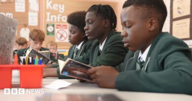 Students in green school uniforms sit at desks reading books during a classroom lesson. Pencil pots and study materials are visible on the tables, with a noticeboard and classroom displays in the background.