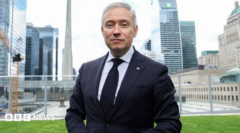 Champagne wears a suit and tie while he stands on a lawn in front of the Toronto city skyline of high-rise buildings.