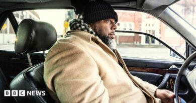 A bearded man in a beige coat and a black and white chequered scarf drives his car through a US urban area. His car has a black leather interior.