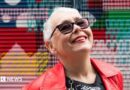 A tightly cropped photo of a woman in her 70s with short grey hair looking up at the sky, smiling. She has sunglasses on, and bright red lipstick that matches her leather jacket. Graffiti is in the background, slightly out of focus.