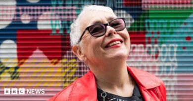 A tightly cropped photo of a woman in her 70s with short grey hair looking up at the sky, smiling. She has sunglasses on, and bright red lipstick that matches her leather jacket. Graffiti is in the background, slightly out of focus.