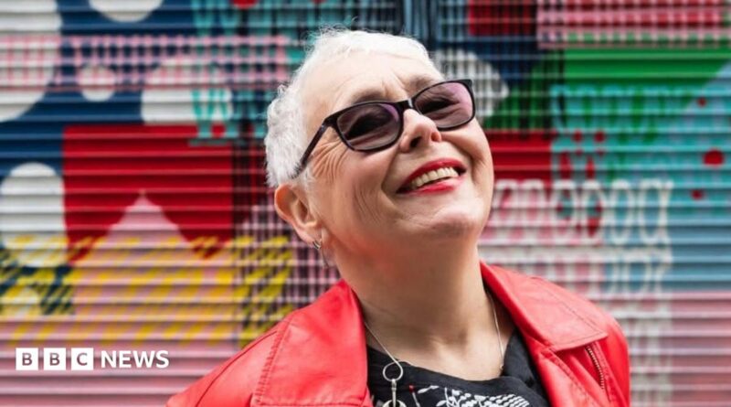 A tightly cropped photo of a woman in her 70s with short grey hair looking up at the sky, smiling. She has sunglasses on, and bright red lipstick that matches her leather jacket. Graffiti is in the background, slightly out of focus.