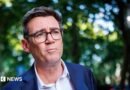 A medium close up of Andy Burnham wearing a blue suit and an open-collar white shirt with trees behind him in soft focus
