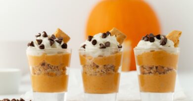 Three shot glasses filled with layers of healthy fall recipes, featuring pumpkin mousse and crushed graham crackers, topped with whipped cream, chocolate chips, and a piece of graham cracker, with a pumpkin in the background.