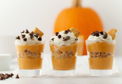 Three shot glasses filled with layers of healthy fall recipes, featuring pumpkin mousse and crushed graham crackers, topped with whipped cream, chocolate chips, and a piece of graham cracker, with a pumpkin in the background.