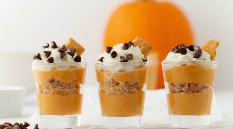 Three shot glasses filled with layers of healthy fall recipes, featuring pumpkin mousse and crushed graham crackers, topped with whipped cream, chocolate chips, and a piece of graham cracker, with a pumpkin in the background.