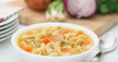 A white bowl of chicken noodle soup with carrots, next to a spoon and a cutting board with red onions and herbs, perfect for healthy soup recipes.