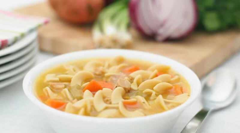 A white bowl of chicken noodle soup with carrots, next to a spoon and a cutting board with red onions and herbs, perfect for healthy soup recipes.