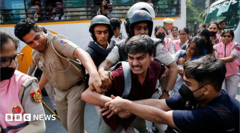 A man is being held by men in official uniforms as another man grapples with them. More people are stood behind in pink uniforms holding other protesters.