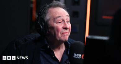 A man with grey hair and wearing a large set of headphones sits behind a BBC Radio 2 microphone.