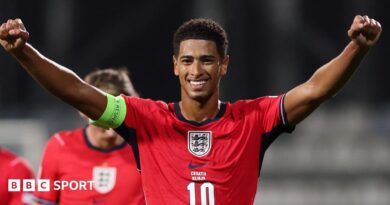 A smiling Jude Bellingham, in red kit, celebrates during England's win over Croatia with both arms stretched out wide to head height