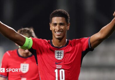 A smiling Jude Bellingham, in red kit, celebrates during England's win over Croatia with both arms stretched out wide to head height
