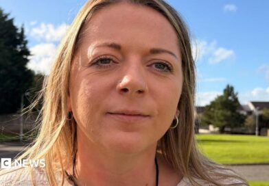 Close-up portrait of a woman with a nose ring and hoop earrings standing outdoors on a sunny day, with a grassy area, trees and houses blurred in the background.