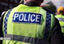 A police officer walks in a crowd alongside other officers. He has an ear piece and is wearing a fluorescent yellow tabard with Police written on the back