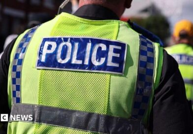 A police officer walks in a crowd alongside other officers. He has an ear piece and is wearing a fluorescent yellow tabard with Police written on the back