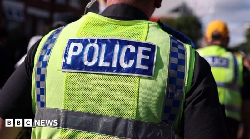 A police officer walks in a crowd alongside other officers. He has an ear piece and is wearing a fluorescent yellow tabard with Police written on the back
