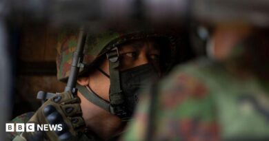 A soldier wearing a helmet and face mask looks to the side, his gloved hand holding the barrel of a rifle, his face only partially visible over the shoulder of another soldier.