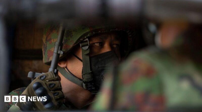 A soldier wearing a helmet and face mask looks to the side, his gloved hand holding the barrel of a rifle, his face only partially visible over the shoulder of another soldier.