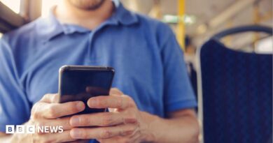 A man in a blue Tshirt holding a mobile phone and sitting on a bus