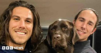 A young man with shoulder-length brown hair and light stubble smiling at the camera, and another young man to his right and a brown dog in between them