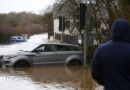 People look on at the cars stuck in the flooding in the hamlet of Weycroft, on January 27, 2026 in Axminster, England.