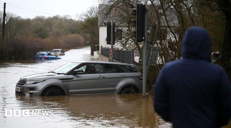 People look on at the cars stuck in the flooding in the hamlet of Weycroft, on January 27, 2026 in Axminster, England.