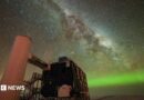 A starry sky the Milky Way is seen through the middle, and the Northern Lights in green along the horizon. In the foreground is an observatory with a black building and stairs liking to a white column. 
