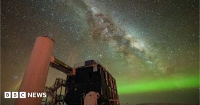 A starry sky the Milky Way is seen through the middle, and the Northern Lights in green along the horizon. In the foreground is an observatory with a black building and stairs liking to a white column. 