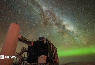 A starry sky the Milky Way is seen through the middle, and the Northern Lights in green along the horizon. In the foreground is an observatory with a black building and stairs liking to a white column. 