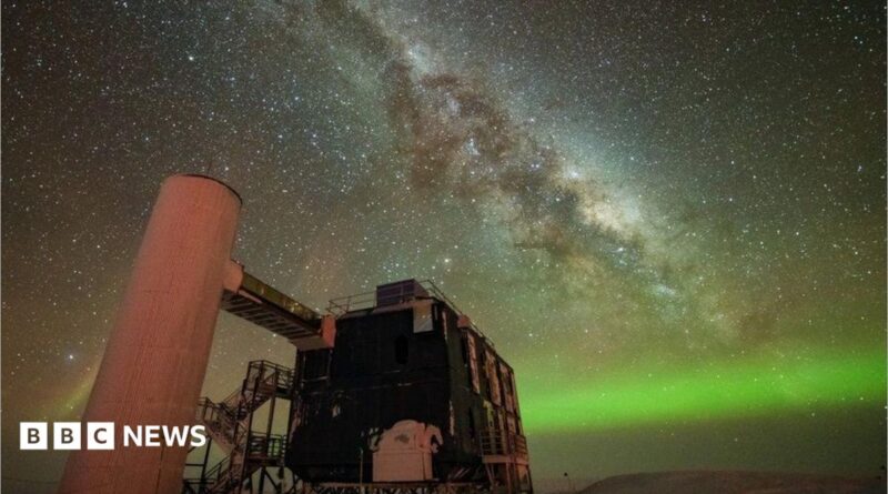 A starry sky the Milky Way is seen through the middle, and the Northern Lights in green along the horizon. In the foreground is an observatory with a black building and stairs liking to a white column.